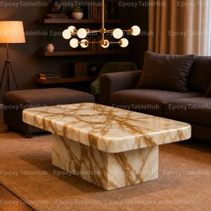 May include: A rectangular coffee table featuring a polished white and gold marble surface with prominent veining. The table has a thick, solid block base made of the same marble material, creating a cohesive and substantial appearance in the room.