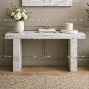 May include: A white marble console table with a rectangular top and two thick, square legs. The table is decorated with a white vase of flowers, a stack of books, a gold sculpture, and a candle on a marble coaster. The table is 122 cm long.
