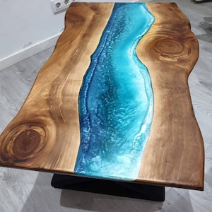 Op de afbeelding: Een houten salontafel met een blauwe en groene epoxyhars rivier die door het midden loopt. De tafel heeft een natuurlijke houten afwerking en een zwarte metalen basis.