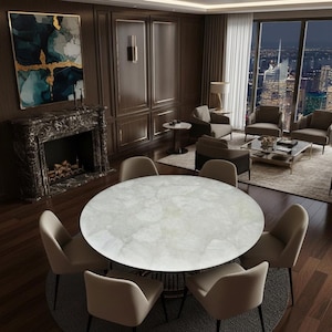 Op de afbeelding: Een luxe eetkamer met een grote, ronde tafel met een lichtgekleurd, patroon oppervlak. Beige stoelen omringen de tafel. De kamer omvat een open haard, kunstwerken en een uitzicht op de stad door grote ramen.