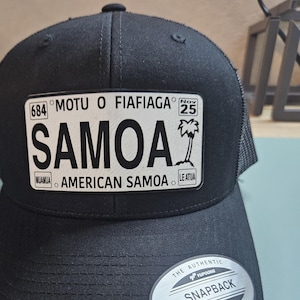 May include: Black snapback hat featuring a rectangular patch with the word "SAMOA" in large white letters. The patch also includes text that reads "AMERICAN SAMOA" and "MOTU O FIAFIAGA". A palm tree graphic is also visible.