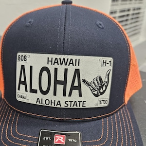 Könnte beinhalten: Eine schwarze und orangefarbene Trucker-Kappe mit einem weißen Patch, auf dem "HAWAII ALOHA" mit einer Handgeste und dem Text "OHANA" und "TATTOO" steht. Die Kappe hat einen schwarzen verstellbaren Riemen mit einem roten "R" und dem Text "RICHARDSON ORIGINAL 112 TRUCKER ADJUSTABLE".