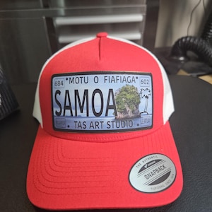 May include: A red and white Samoa trucker hat with a rectangular patch featuring the text "SAMOA" and "TAS ART STUDIO". The patch also includes an image of a tropical scene. The hat has a snapback closure.