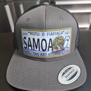 Op de afbeelding: Grijze en zwarte snapback cap met een rechthoekige patch met het woord "SAMOA". De patch bevat een afbeelding van een tropisch eiland en de tekst "MOTU O FIAFIAGA" en "TAS ART STUDIO".
