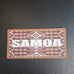 May include: A rectangular brown coaster with the word "SAMOA" in white block letters. The coaster has a brown border and a detailed geometric pattern in shades of brown and white.