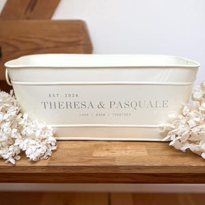 Könnte beinhalten: Cremefarbener rechteckiger Metallpflanzkübel mit dem Text "THERESA & PASQUALE" und "LOVE | GROW | TOGETHER". Der Pflanzkübel steht auf einer Holzoberfläche, mit weißen Trockenblumen. Der Text "EST. 2026" steht über den Namen.