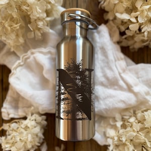 Könnte beinhalten: Eine Edelstahl-Wasserflasche mit Bambusdeckel. Die Flasche hat eine schwarze Grafik eines Baumes und das Wort "Vibbe" in Handschrift. Die Flasche ist von weißen Blumen und einem weißen Tuch umgeben. Höhe ca. 25 cm.