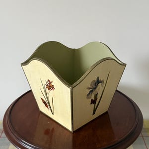 May include: A cream-coloured wooden container with a scalloped rim, decorated with floral illustrations and gold trim. It sits on a dark brown, round table. The interior of the container is light green.