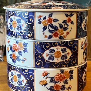 May include: A three-tiered, lidded ceramic container with a traditional Japanese design. The container features alternating panels of white and deep blue, adorned with floral and swirling patterns in orange, yellow, and gold. The stacked design is ideal for serving or storage.