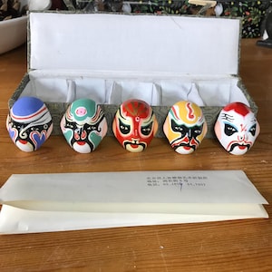 May include: Five small, colorful, decorative Chinese opera masks in a display box. Each mask features a unique design with vibrant colors like red, blue, yellow, and green. The masks are arranged in a row, showcasing intricate details.