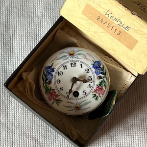Puede incluir: Una esfera de reloj redonda vintage con fondo blanco, números negros y delicados diseños florales en rosa, azul y amarillo. El reloj está en una caja negra con tapa beige, y el texto "KEINZLE 24/5113" es visible en una etiqueta.