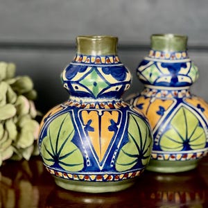 Könnte beinhalten: Zwei dekorative Keramikvasen mit bauchiger Form und aufwendigen handgemalten Designs. Die Vasen haben eine Farbpalette aus Blau, Orange, Grün und Rot mit einem grünen Rand. Das Design umfasst Blatt- und geometrische Muster. Die Vasen stehen auf einer dunklen Holzoberfläche.