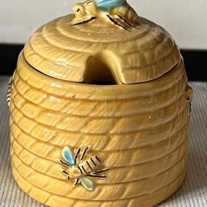 May include: A ceramic honey pot shaped like a beehive, with a lid and a small opening for a spoon. The pot is yellow with a textured surface, and a bee figurine is on top. A bee is also painted on the side. The pot is a decorative kitchen item.