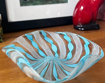 Könnte beinhalten: Eine dekorative Glasschale mit quadratischer Form und Wirbeldesign. Die Schale hat abwechselnde Bänder in Hellblau, Weiß und Braun. Die Schale steht auf einer Holzoberfläche.