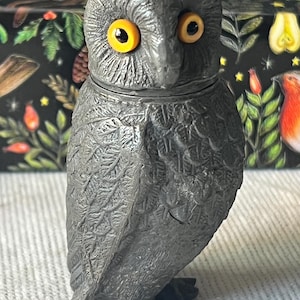 Pode incluir: Estatueta de coruja cinza escuro detalhada com olhos amarelo brilhante. A coruja tem penas texturizadas e uma cabeça pequena e redonda. A estatueta está em pé, com os pés visíveis. O fundo é uma superfície escura com um design floral colorido.