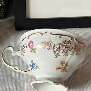 以下が含まれることがあります： ピンク、青、黄色などの花柄が施された、金色の取っ手と縁が付いた白い磁器のティーカップ。カップはスカラップのベースと装飾的な金のアクセントが特徴で、ヴィンテージまたはアンティークスタイルを示唆しています。