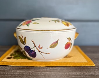 May include: A white ceramic serving bowl with a lid, adorned with a fruit and leaf design in shades of red, purple, and yellow. The bowl has gold-colored handles and trim. It sits on a yellow placemat.