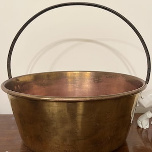 May include: An antique copper cauldron with a dark metal handle. The cauldron has a wide, rounded body and a slightly flared rim. The interior of the cauldron has a reddish hue, while the exterior has a tarnished, aged appearance.