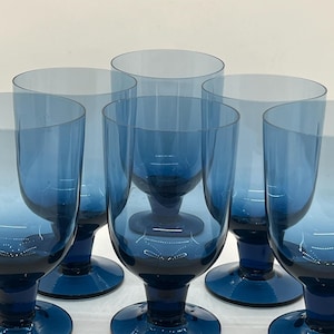 May include: Six blue-tinted glass goblets with a rounded bowl and a short stem. The goblets have a dark blue base that fades to a lighter blue towards the rim. The glasses are arranged in a group, showcasing their design and color.