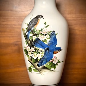 Può includere: Vaso in ceramica bianca con un passero azzurro e un ramo di fiori bianchi. Il vaso ha un bordo dorato e una base in legno. La firma dell'artista, "Roger Tory Peterson", è visibile sul vaso.