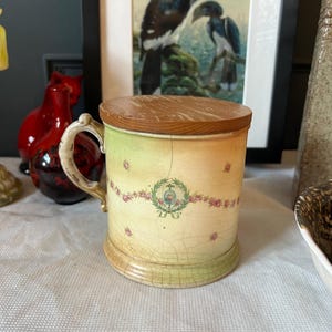 May include: A vintage ceramic mug with a wooden lid. The mug has a cream-coloured base with a floral and crest design. The handle is off-white, and the lid is made of light brown wood. The mug has a crackled appearance.