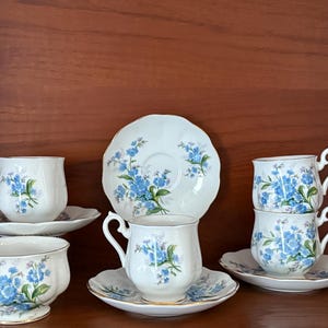 Può includere: Un set di tazze e piattini vintage. Le tazze e i piattini in porcellana bianca sono decorati con motivi floreali blu. Il set comprende sei tazze, sei piattini e un piatto da portata. Le tazze hanno manici curvi.
