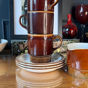 May include: Three brown ceramic mugs stacked, with matching saucers beneath. The mugs and saucers have a glossy finish and a ribbed design at the base. A matching creamer is also visible, all on a wooden surface.