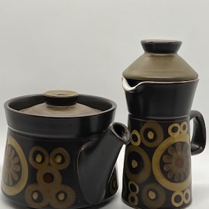 以下が含まれることがあります： ティーポットとコーヒーポットを備えたヴィンテージセラミックティーセット。セットは主に濃い茶色で、黄色と茶色の円と花の模様が繰り返されています。どちらの作品にも茶色の蓋が付いています。
