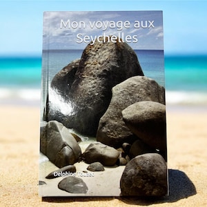 Peut inclure: Couverture de livre "Mon voyage aux Seychelles" avec une scène de plage, de gros rochers gris et l'océan. Le nom de l'auteur, Delphine Touzec, est en bas. En arrière-plan, une plage de sable et de l'eau bleue.