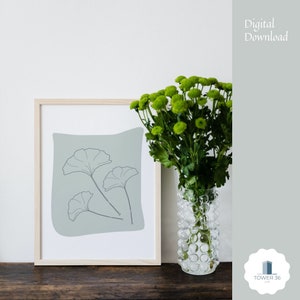 Può includere: Una stampa incorniciata di un disegno di linea minimalista di tre foglie di ginkgo su uno sfondo blu chiaro. La stampa è in una cornice di legno chiaro e si trova su una superficie di legno. Un vaso di fiori verdi è in primo piano.