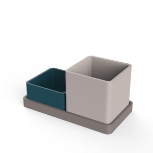 Può includere: Un moderno organizer da scrivania di forma quadrata. È dotato di un contenitore quadrato blu teal, un contenitore quadrato beige più grande e una base rettangolare grigia. L'organizer è progettato per contenere penne, matite e altri articoli per ufficio.