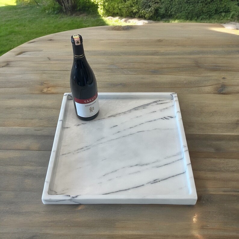 May include: A rectangular white marble tray with gray veining, holding a bottle of red wine. The wine bottle has a black label with white text. The tray is on a wooden surface.