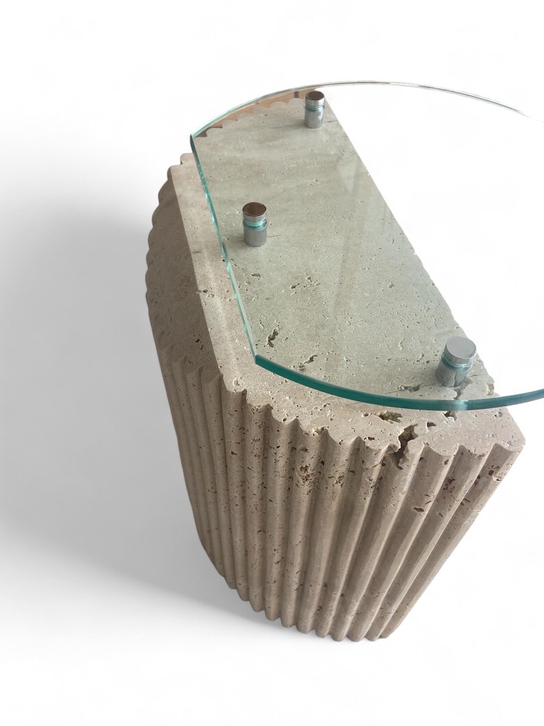 May include: A half-round side table with a fluted travertine base and a clear glass top. The table has three silver metal accents on the top.