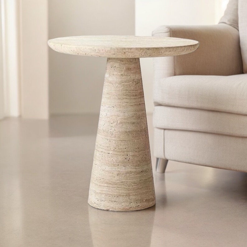 May include: A beige, round side table with a conical base. The table is made of a stone-like material and has a diameter of approximately 20 inches. The table is a neutral color and would complement a variety of decor styles.