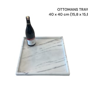 May include: A square marble-look ottoman tray, 15.8 x 15.8 inches, with a raised edge. A bottle of red wine rests on the tray. The tray has a white and gray veined pattern, and the text "OTTOMANS TRAY" is visible.