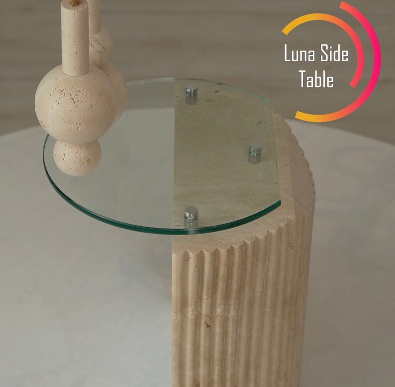 May include: A beige travertine side table with a round glass top. The table has a fluted design and the glass top has a rounded edge.