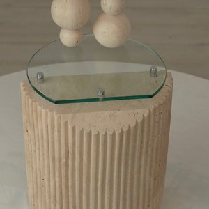 May include: A beige, textured, cylindrical pedestal with a round glass top. The pedestal has a fluted design and the glass top has a rounded edge.