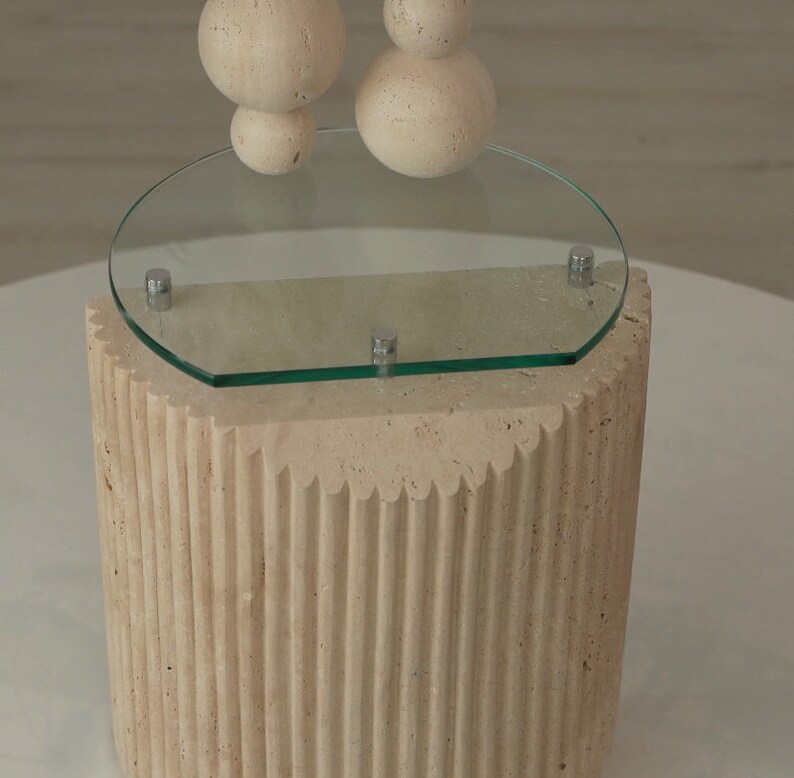 May include: A beige, textured, cylindrical pedestal with a round glass top. The pedestal has a fluted design and the glass top has a rounded edge.