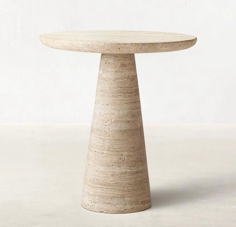 May include: A beige, round side table with a conical base. The table is made of a light-colored stone material, with a smooth, circular top. The base tapers from a wider bottom to a narrower top, creating a visually appealing design. The table is approximately 24 inches tall.