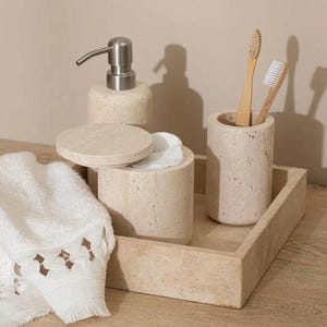 Peut inclure: Un plateau de salle de bain en travertin beige avec un distributeur de savon, un récipient pour cotons-tiges, un porte-brosses à dents et une serviette blanche. Le plateau est en pierre naturelle et a un aspect rustique.