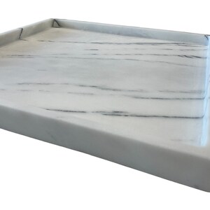 May include: A rectangular white marble tray with gray veining. The tray has a raised edge and a smooth, polished surface. The corner of the tray is visible, showing the clean lines and the quality of the stone.
