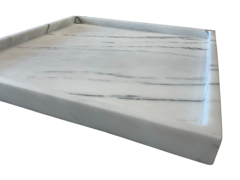May include: A rectangular white marble tray with gray veining. The tray has a raised edge and a smooth, polished surface. The corner of the tray is visible, showing the clean lines and the quality of the stone.