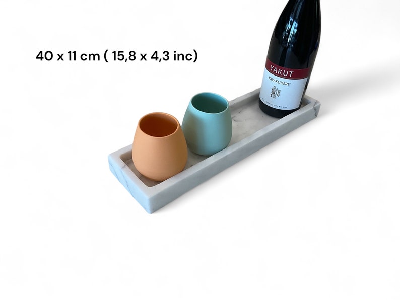 May include: A rectangular marble tray with a bottle of red wine and two ceramic cups. The tray measures 15.8 x 4.3 inches. One cup is peach-colored, and the other is light blue. The wine bottle has a white label with red text.