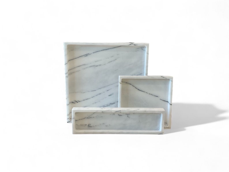 May include: Three white marble trays of varying sizes. The trays have a rectangular shape with slightly raised edges. Each tray features gray veining patterns throughout the marble, adding visual interest. The trays are arranged on a white surface.