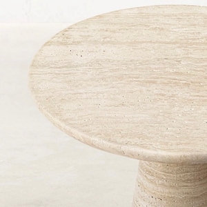 May include: A round, beige stone table with a textured surface. The table has a single, cylindrical base. The tabletop is approximately 30 inches in diameter. The table is made of a light-colored stone.