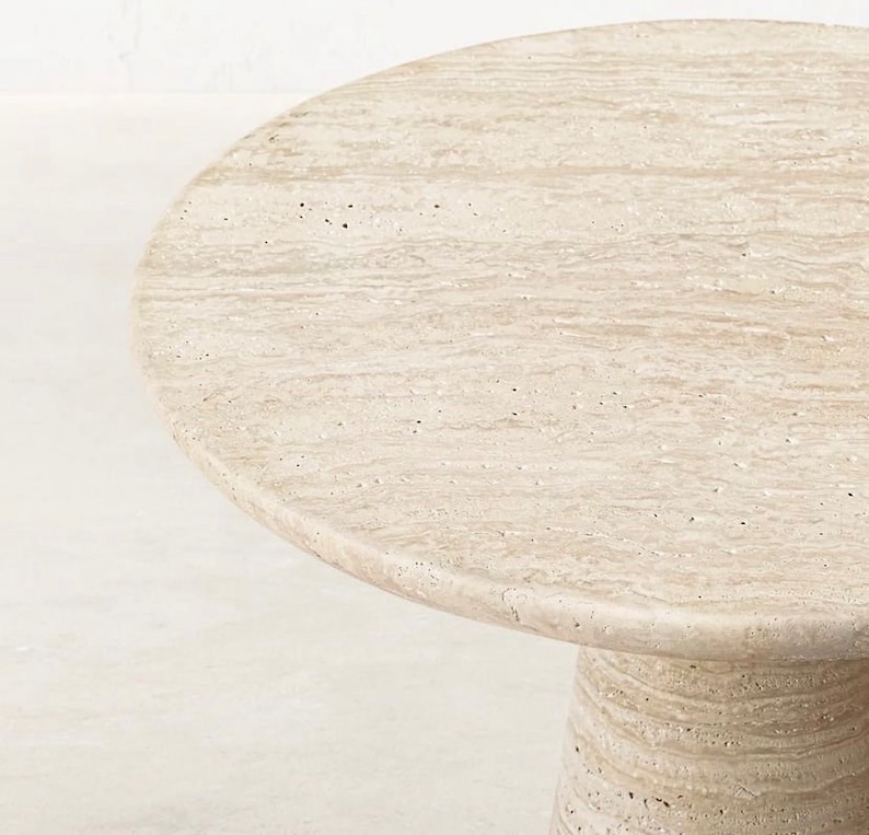 May include: A round, beige stone table with a textured surface. The table has a single, cylindrical base. The tabletop is approximately 30 inches in diameter. The table is made of a light-colored stone.