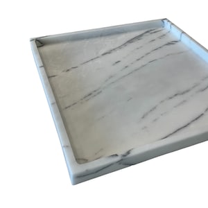 May include: A rectangular white marble tray with dark gray veining. The tray has a raised edge and a smooth surface. The marble pattern is consistent across the tray's surface, creating a cohesive design. The tray is suitable for serving or display.