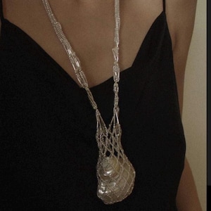 Op de afbeelding: Een zilveren ketting met een schelpvormige hanger. De hanger is gemaakt van een fijn, geweven gaas en hangt aan een lange, zilveren ketting.