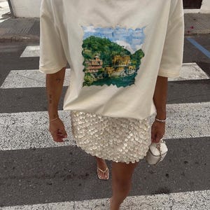 May include: Cream-colored t-shirt with a scenic print of a coastal town, paired with a sequined mini-skirt. The outfit is completed with gold sandals and a small white handbag. The image is taken on a crosswalk.