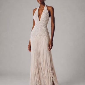May include: A floor-length, off-white halter dress featuring a deep V-neckline. The bodice is densely covered in small, textured beads, while the skirt consists of long, flowing fringe that extends to the hem, creating a layered, linear appearance.